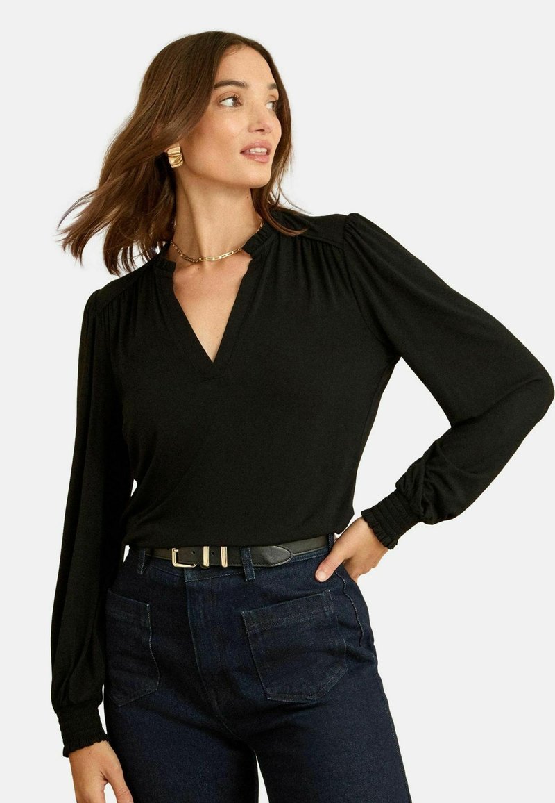 Fekete, hosszú ujjú blúz V kivágással, puffos vállrészekkel és kiszűkített mandzsettákkal, sötét farmernadrággal és fekete övvel párosítva.