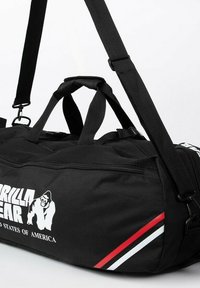 Black duffel bag with two top handles and a detachable shoulder strap. Features a white logo and red, white, and green stripes. Textured fabric.