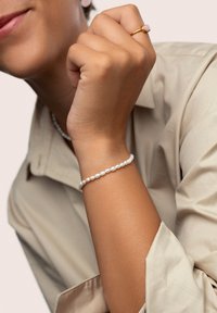 Perlenarmband mit kleinen, glatten weißen Perlen; Hand, die einen goldenen Ring mit einem rosa Stein trägt; Oberteil in neutralen Farbtönen; Nahaufnahme mit Fokus auf die Accessoires.