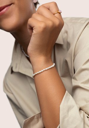 Perlenarmband mit kleinen, glatten weißen Perlen; Hand, die einen goldenen Ring mit einem rosa Stein trägt; Oberteil in neutralen Farbtönen; Nahaufnahme mit Fokus auf die Accessoires.
