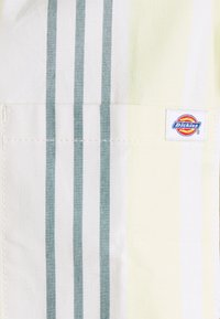 Randigt arbetsklädesmaterial med vertikala ljusgula och gröna ränder samt en framträdande blå Dickies-logotyp. Slät textur.