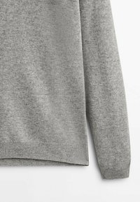 Gros plan sur une manche et un ourlet d'un pull en tricot gris clair avec des poignets et un bord inférieur côtelés sur un fond blanc.