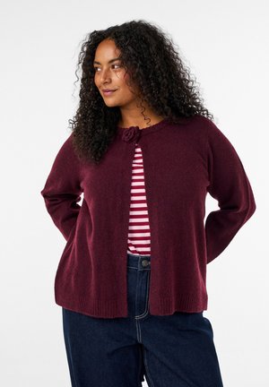 Femme aux cheveux bouclés portant un cardigan bordeaux sur une chemise rayée rouge et blanche et un jean bleu foncé, regardant sur le côté avec les mains derrière le dos.
