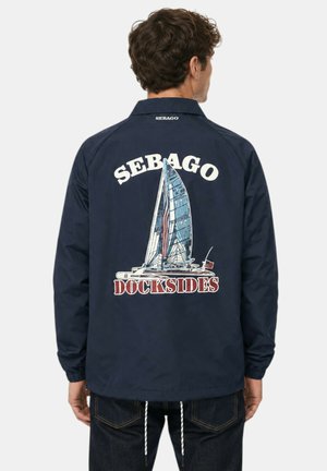 Uomo che indossa una giacca blu navy con una grande grafica di una barca a vela e il testo "Sebago Dockside" sulla schiena, in piedi di spalle su sfondo semplice.
