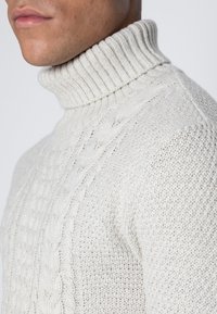 Krämfärgad polotröja i texturerad stickad kvalitet. Har ribbat krage, kabel- och bikakmönster. Tätt sittande design.