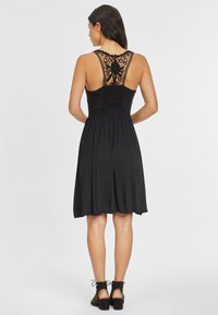 Schwarzes Kleid mit engem Oberteil und fließendem Rock. Auffällige Spitzenverzierungen am Rücken, verstellbare Träger und eine weiche Textur.