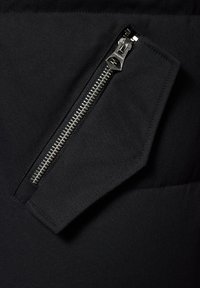 Czarna tkanina o teksturowanym wykończeniu posiada ukośną kieszeń na zamek błyskawiczny z srebrnym okuciem, co podkreśla funkcjonalność i detale projektu.