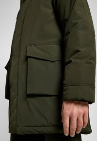 Oliwkowa kurtka puchowa z kieszenią na piersi, teksturowanym materiałem i luźnym krojem. Widoczne detale szwów podkreślają design.