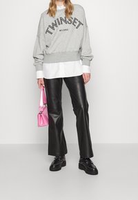 Sweatshirt gris avec le logo "TWINGSET MILANO", superposé à une chemise blanche. Un pantalon en cuir noir évasé et un sac à main en vernis rose complètent le look.
