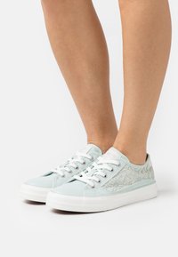 Světle modré plátěné tenisky s tkaničkovou panele, bílé gumové podrážky a ploché bílé tkaničky. Vyznačují se nízkým střihem a texturovaným svrškem.