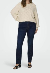 Beige stickad tröja med diamantmönster och små dekorationer, tillsammans med mörkblå denimjeans och svarta blockklacksandaler.