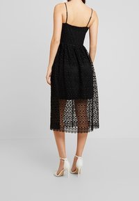 Svart spetsklänning med en åtsittande bodice och full, halvgenomskinlig kjol. Har tunna axelremmar och en vågig fåll. Bärs med vita klacksandaler.