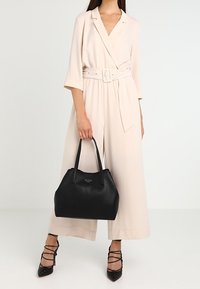 Femme portant une combinaison crème ceinturée et tenant un sac à main en cuir noir, vêtue de talons aiguilles noirs à lanières, se tenant contre un fond uni.