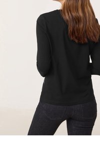Donna con lunghi capelli castani, che indossa un top nero a maniche lunghe e jeans blu scuro, vista da dietro su uno sfondo semplice.