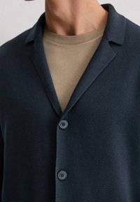 Dunkelblauer Strickcardigan mit Kragen, ausgestattet mit zwei Knöpfen und einem strukturierten Stoff. Getragen über einem hellbraunen Rundhals-Shirt.