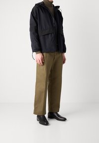 Černá lehká bunda s kapucí, se zipem vpředu a elastickými manžetami; kombinovaná se zelenými kalhotami olivové barvy a černými kotníkovými botami.