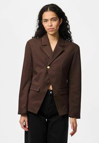 Blazer marron ajusté avec un revers cranté, fermeture à un bouton doré, et deux poches frontales ; porté sur un pantalon noir taille haute.