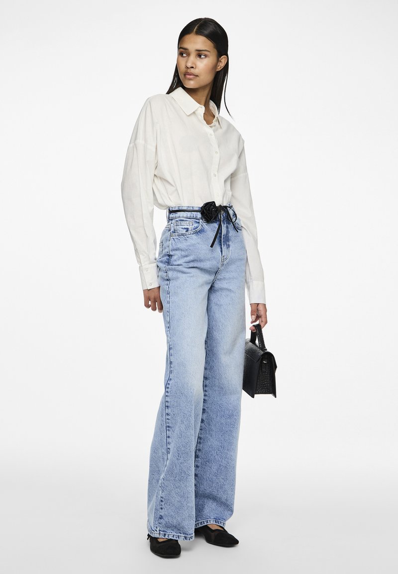 Witte lange mouwen overhemd met knopen, hoog gewaisted lichtblauwe wijde jeans, zwarte rozenriem, zwarte handtas en zwarte flats.
