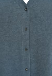 V-kaula, napapa shirt tummansinisenvihreä kangas jolla on teksturoitu pinta. Edessä on viisi tummaa nappia ja kapeat tikit.