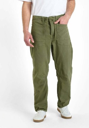 Man in olijfgroene cargobroek met grote voorkruikezakken en een strikceintuur, gecombineerd met witte sneakers en een wit shirt.