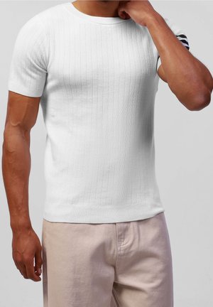 Pull en tricot côtelé blanc à manches courtes et col rond. Agrémenté d'un détail à rayures noires sur la manche. Porté avec un pantalon de couleur claire.