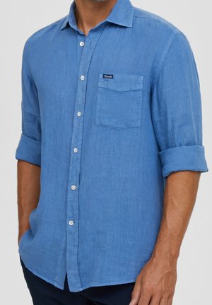 Mann trägt blaues langärmeliges Hemd mit Brusttasche und hochgekrempelten Ärmeln, Hände in den Taschen, vor einfarbigem Hintergrund.