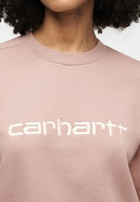 Φούτερ σε σκονισμένο ροζ με απαλό ύφασμα, στρογγυλή λαιμόκοψη και έντονο λευκό κεντητό λογότυπο "carhartt" στο στήθος.
