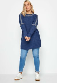 Blauer gestrickter Pullover mit rundem Ausschnitt und dekorativen Details an den Ärmeln, kombiniert mit hellblauen Jeans und weißen Sneakers.