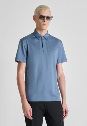 Jonge man met kort blond haar, donkere zonnebril, een blauwe polo met korte mouwen, zwarte broek en een kleine tattoo op zijn linkerarm.
