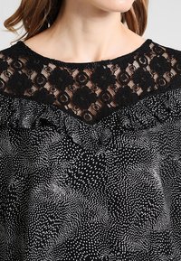 Blusa nera con scollatura in pizzo a motivo floreale, bordi arricciati e un design a pois bianchi su tutta la stoffa.