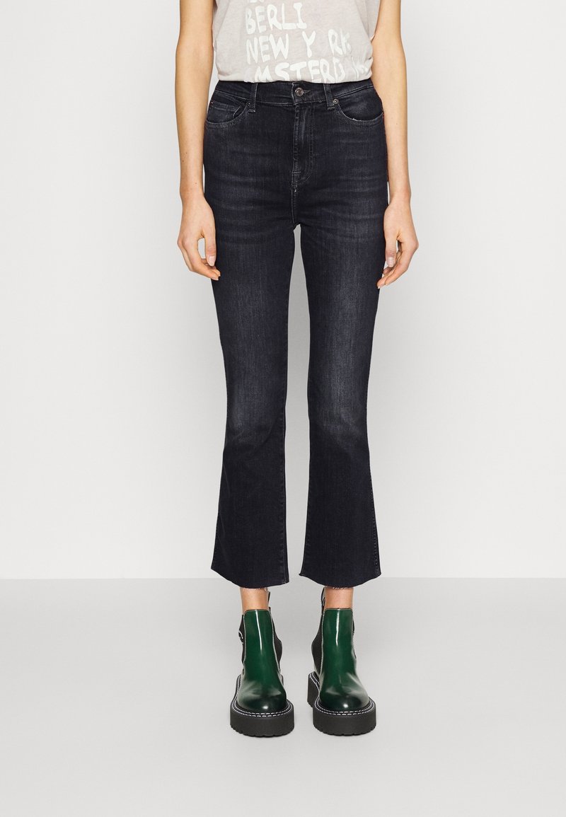 Personne portant un jean noir flare court taille haute et des bottines vert foncé brillantes à semelles épaisses sur un fond uni.