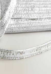 Sølv håndtaske med en tekstureret, metallisk finish. Har en glat, flad rem, der reflekterer lys. Detaljer inkluderer et vævet mønster.
