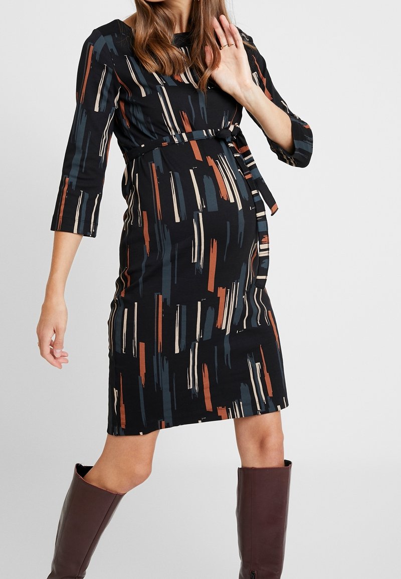 Femme enceinte portant une robe noire à motifs longueur genou avec manches 3/4 et des bottes marron montantes au genou, posant avec une main levée.