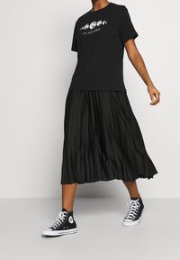 Camiseta negra con gráfico de fases lunares y texto, combinada con una falda midi plisada negra y zapatillas altas negras.