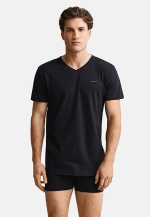 Hombre de pie con camiseta negra de cuello en V y calzoncillos tipo bóxer negros contra un fondo blanco liso.