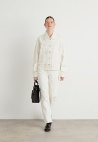 Hvit denimjakke med snor i livet, knappelukking foran, og to lommer foran, kombinert med hvite bukser og en liten svart veske.