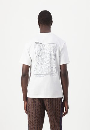 Homme portant un t-shirt blanc avec un motif de carte contour noir dans le dos et un pantalon marron à motifs, debout sur un fond blanc uni.