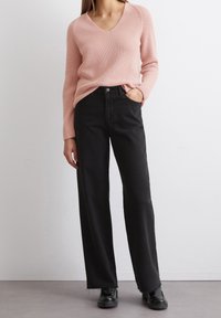 Pull en maille côtelée rose avec un col en V, associé à un jean noir taille haute avec une coupe droite et un ourlet légèrement effiloché. Chaussures noires.