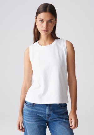 Donna con capelli castani lisci che indossa una canotta bianca e jeans blu, in piedi davanti a uno sfondo chiaro e uniforme.