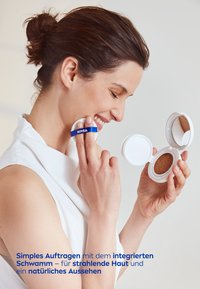 Weißer kompakter Spiegel, der einen braunen Schwamm enthält. Benutzer trägt das Produkt mit einem angebrachten blauen Gurt mit der Aufschrift "NIVEA" auf das Gesicht auf. Hellblauer Hintergrund.