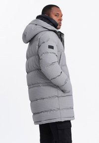 Giacca puffer grigia con motivo a quadri, dotata di un cappuccio nero, tasche laterali e un'etichetta del marchio sulla manica sinistra.