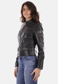Giacca di pelle nera con design aderente, colletto alto, zip frontale e due tasche anteriori. Texture liscia con un leggero luccichio.