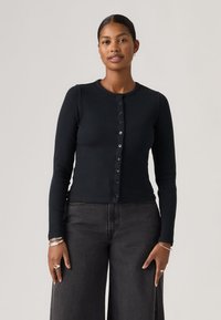 Cárdigan negro de canalé con escote redondo, mangas largas y cierre frontal con botones. La modelo lo lleva combinado con jeans oscuros y holgados.