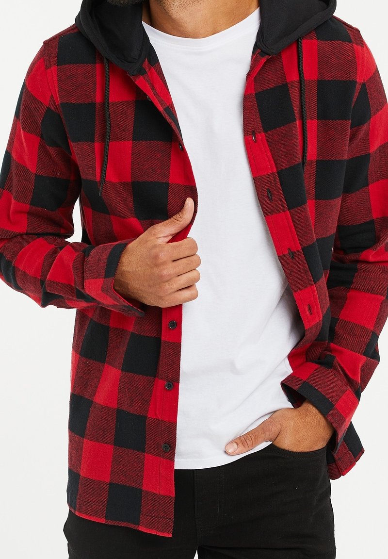 Man met een rood en zwart geblokt hooded shirt over een wit T-shirt, waarbij hij met één hand het shirt aanpast en de andere in zijn zak heeft.