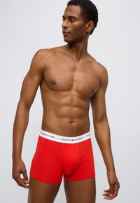 Κόκκινα βαμβακερά ανδρικά εσώρουχα σε στυλ μποξέρ με λευκή ελαστική μέση που φέρει λογότυπο. Η εφαρμογή είναι στενή και το ύφασμα έχει λεία υφή.