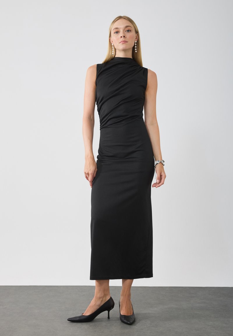 Abito nero senza maniche con dettagli arricciati, scollatura alta e design lungo fino al pavimento, abbinato a décolleté nere a punta.