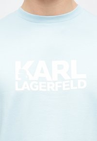 Világoskék pamutpóló kerek nyakkivágással, elöl vastag fehér „KARL LAGERFELD” felirattal, enyhe textúrával.