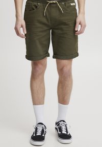 Olívazöld rövidnadrág húzózsinóros derékrésszel, felhajtott szegélyekkel és két első zsebbel, fehér zoknival és fekete sportcipővel párosítva.