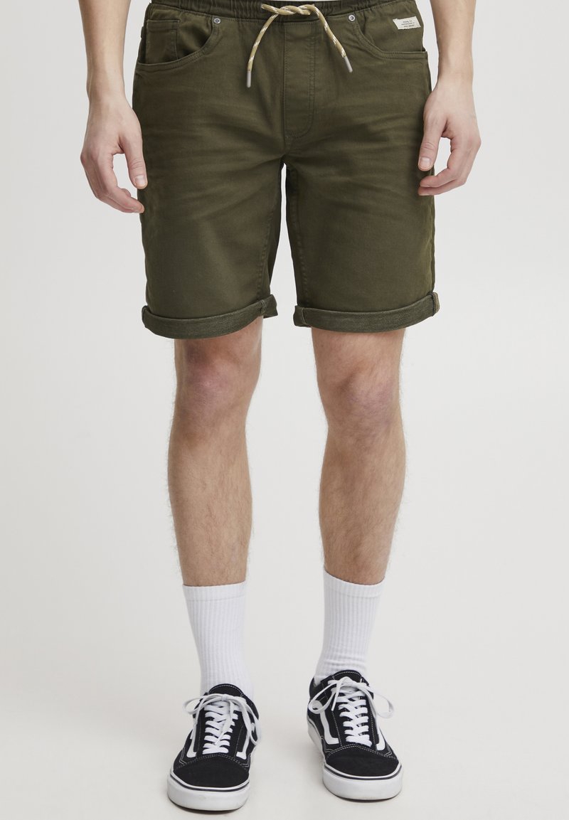 Olívazöld rövidnadrág húzózsinóros derékrésszel, felhajtott szegélyekkel és két első zsebbel, fehér zoknival és fekete sportcipővel párosítva.