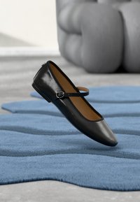 Scarpa piatta in pelle nera con punta appuntita e una sottile fascia con fibbia. Sullo sfondo un tappeto blu con trama.
